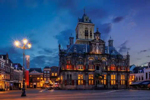 Fachada iluminada del ayuntamiento histórico de Delft, con su torre, en la plaza del mercado al anochecer