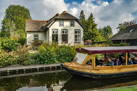 Barca turística de madera con toldo pasando junto a una casa de ladrillo blanco con tejado de junco en un canal de Giethoorn