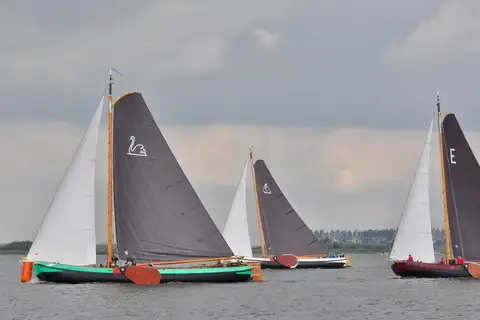 Tres veleros tradicionales frisones con velas oscuras compitiendo en un lago de Frisia bajo un cielo nublado
