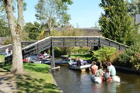 Barcas con familias pasando bajo un puente de madera en un canal de Giethoorn, entre jardines y casas de ladrillo