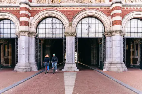 Fachada del Rijksmuseum con sus arcos de ladrillo y piedra y dos ciclistas saliendo del paso bajo el edificio