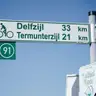 Cartel verde de ruta ciclista en neerlandés indicando Delfzijl y Termunterzijl con el número de ruta 91 bajo un cielo azul