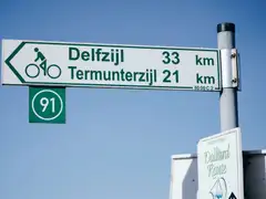 Cartel verde de ruta ciclista en neerlandés indicando Delfzijl y Termunterzijl con el número de ruta 91 bajo un cielo azul