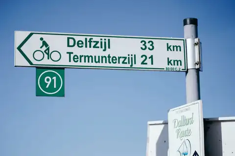 Cartel verde de ruta ciclista en neerlandés indicando Delfzijl y Termunterzijl con el número de ruta 91 bajo un cielo azul