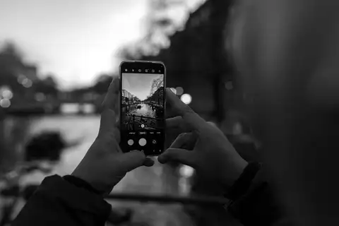 Manos sosteniendo un teléfono móvil cuya pantalla muestra un canal de Ámsterdam al anochecer, en blanco y negro