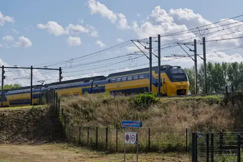 Tren de dos pisos amarillo y azul de NS circulando junto a un talud con hierba y catenaria bajo un cielo con nubes