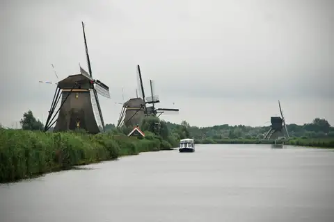 Molinos de Kinderdijk a la orilla del canal, con un barco turístico pequeño navegando por el agua bajo cielo gris