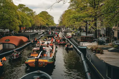 Barcos pequeños llenos de gente con detalles naranjas navegando por un canal arbolado de Ámsterdam, entre casas flotantes