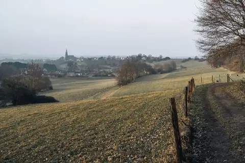 Ladera de campos otoñales con un camino y una valla de madera, y al fondo un pueblo con el campanario de su iglesia, cerca de Heerlen