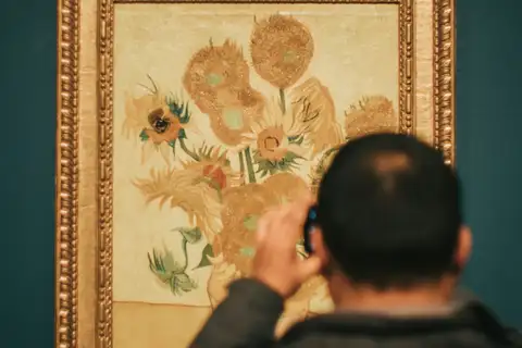 Visitante de espaldas fotografiando Los girasoles de Van Gogh, con su marco dorado sobre una pared verde en el Museo Van Gogh
