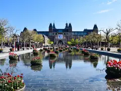 Rijksmuseum al fondo de la Museumplein, con un estanque que refleja el edificio, maceteros de tulipanes y visitantes paseando