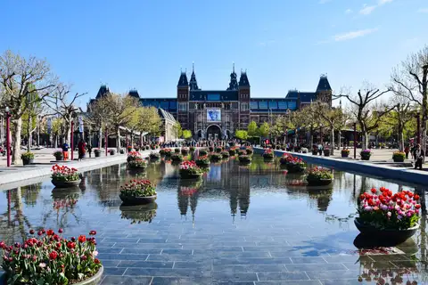 Rijksmuseum al fondo de la Museumplein, con un estanque que refleja el edificio, maceteros de tulipanes y visitantes paseando