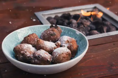 Oliebollen holandeses espolvoreados con azúcar glas en un cuenco azul junto a un brasero encendido
