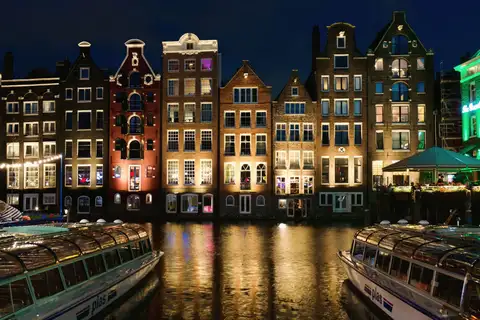Barcos turísticos de techo acristalado amarrados de noche ante casas de canal iluminadas que se reflejan en el agua de Ámsterdam