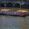 Barco turístico de techo acristalado y detalles naranjas navegando por un canal de Ámsterdam ante un puente de arcos de ladrillo