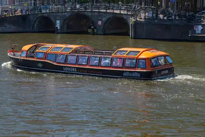 Barco turístico de techo acristalado y detalles naranjas navegando por un canal de Ámsterdam ante un puente de arcos de ladrillo