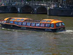 Barco turístico de techo acristalado y detalles naranjas navegando por un canal de Ámsterdam ante un puente de arcos de ladrillo