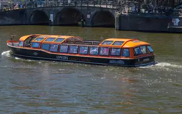 Barco turístico de techo acristalado y detalles naranjas navegando por un canal de Ámsterdam ante un puente de arcos de ladrillo