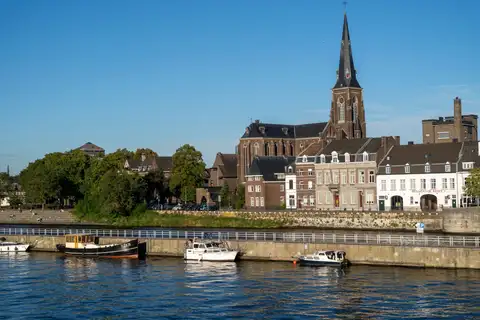 Orilla del Mosa en Maastricht con casas históricas, una iglesia de aguja oscura y barcos amarrados bajo cielo azul