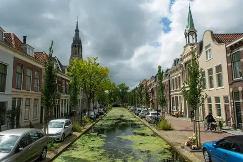 Canal de Delft cubierto de lentejas de agua entre casas históricas de ladrillo y árboles, con una torre de iglesia