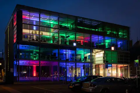 Edificio de oficinas de cristal iluminado con luces de colores verdes, azules y rojas en Eindhoven al anochecer