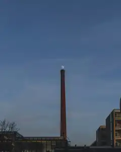 Alta chimenea de ladrillo entre edificios industriales y modernos de Eindhoven al anochecer, con un cielo azul profundo