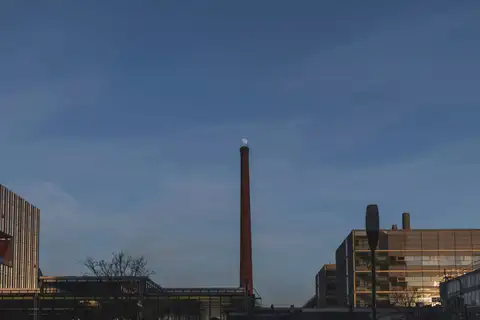 Alta chimenea de ladrillo entre edificios industriales y modernos de Eindhoven al anochecer, con un cielo azul profundo