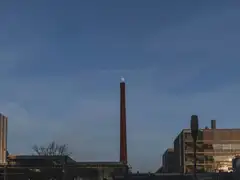 Alta chimenea de ladrillo entre edificios industriales y modernos de Eindhoven al anochecer, con un cielo azul profundo