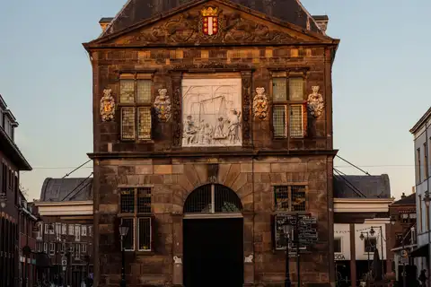 Edificio histórico de piedra y ladrillo con frontón triangular y gran puerta central, iluminado por el sol del atardecer