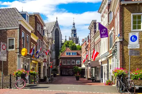 Calle de Gouda con casas de ladrillo, banderas, flores y bicicletas, y la torre de una iglesia gótica al fondo
