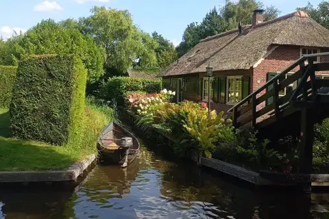 Casa de ladrillo con tejado de junco junto a un canal estrecho de Giethoorn, con una barca amarrada, flores y un puente de madera