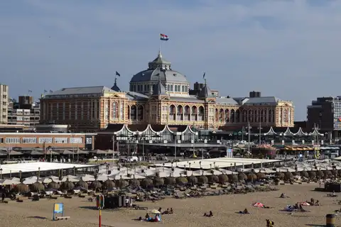 Hotel Kurhaus de ladrillo con cúpula y bandera neerlandesa sobre la playa de Scheveningen, con sombrillas y bañistas en la arena
