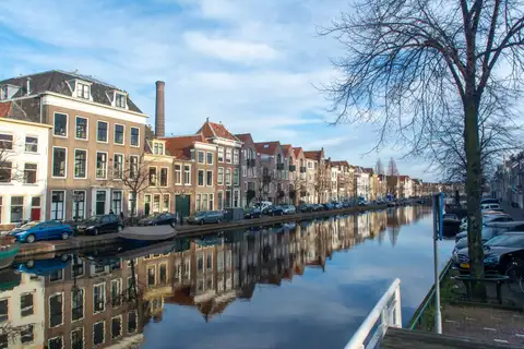 Canal de Leiden con casas de ladrillo del siglo XVII reflejadas en el agua, una chimenea de fábrica al fondo y un árbol sin hojas