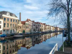 Canal de Leiden con casas de ladrillo del siglo XVII reflejadas en el agua, una chimenea de fábrica al fondo y un árbol sin hojas