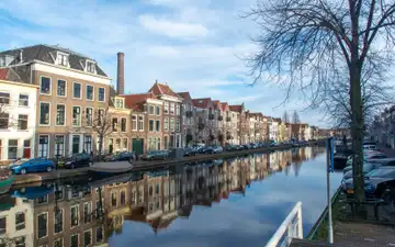 Canal de Leiden con casas de ladrillo del siglo XVII reflejadas en el agua, una chimenea de fábrica al fondo y un árbol sin hojas