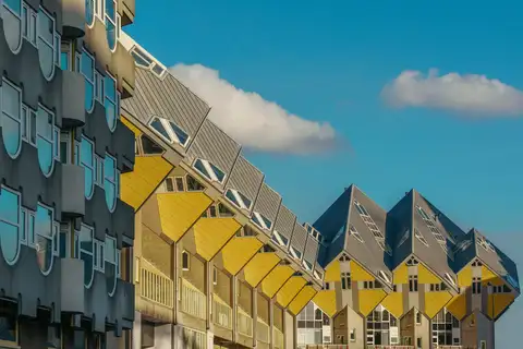 Hilera de casas cubo amarillas inclinadas de Piet Blom junto a un edificio gris de balcones curvos, bajo un cielo azul