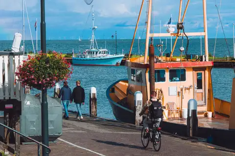 Pesqueros amarillos amarrados en el muelle del puerto de Volendam, con paseantes, un ciclista y una barca azul bajo un cielo despejado
