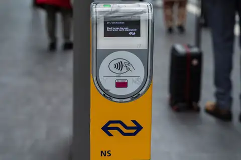 Lector amarillo de NS para pagar con tarjeta sin contacto o OV-chipkaart en un andén de estación holandesa, con viajeros y una maleta desenfocados
