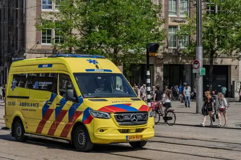 Ambulancia amarilla con franjas rojas y azules circulando por una plaza adoquinada de Ámsterdam entre peatones y ciclistas