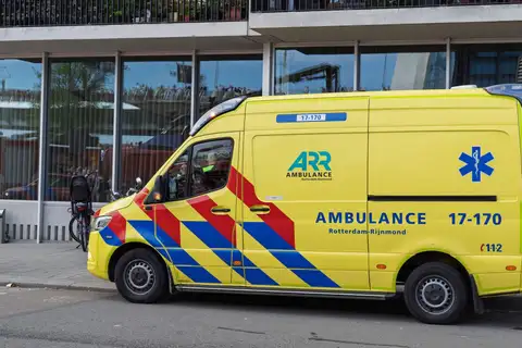 Ambulancia amarilla de Rotterdam-Rijnmond aparcada frente a un edificio de cristal, con una bicicleta con sillita infantil al lado