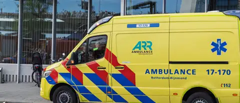 Ambulancia amarilla de Rotterdam-Rijnmond aparcada frente a un edificio de cristal, con una bicicleta con sillita infantil al lado