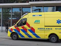 Ambulancia amarilla de Rotterdam-Rijnmond aparcada frente a un edificio de cristal, con una bicicleta con sillita infantil al lado