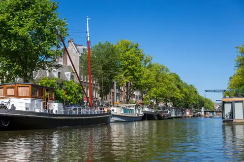 Barcazas y casas flotantes amarradas bajo los árboles en un canal de Ámsterdam, con un puente levadizo al fondo y cielo azul