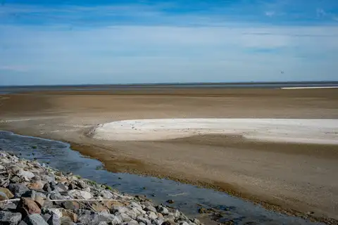 Llanura de arena y lodo del mar de Wadden en marea baja, con un canal de agua junto a una escollera de piedras y cielo azul