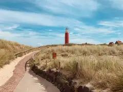 Sendero de ladrillo entre dunas con hierba hacia el faro rojo de Texel, con unas casas al fondo bajo un cielo azul con nubes
