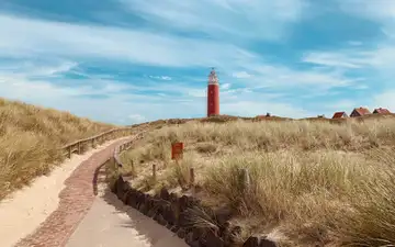 Sendero de ladrillo entre dunas con hierba hacia el faro rojo de Texel, con unas casas al fondo bajo un cielo azul con nubes