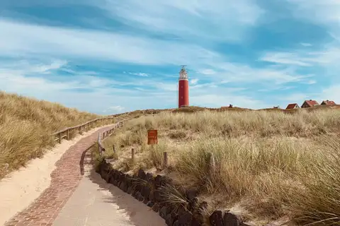 Sendero de ladrillo entre dunas con hierba hacia el faro rojo de Texel, con unas casas al fondo bajo un cielo azul con nubes