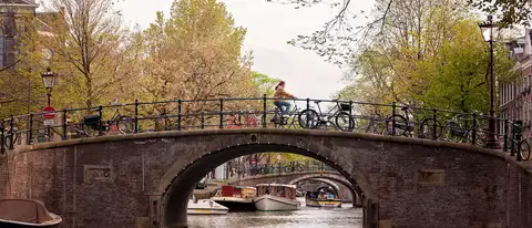 Ciclista cruzando un puente de ladrillo con bicicletas aparcadas sobre un canal de Ámsterdam, con barcos y árboles de primavera alrededor