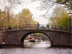 Ciclista cruzando un puente de ladrillo con bicicletas aparcadas sobre un canal de Ámsterdam, con barcos y árboles de primavera alrededor