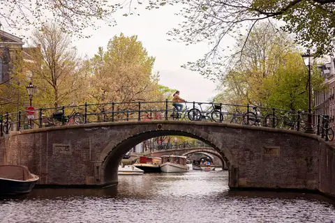Ciclista cruzando un puente de ladrillo con bicicletas aparcadas sobre un canal de Ámsterdam, con barcos y árboles de primavera alrededor